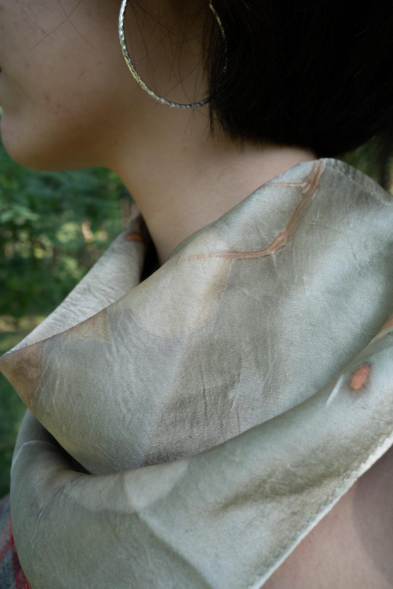 May include: A soft, sage green scarf featuring subtle, organic brown leaf-like imprints and vein patterns across the fabric. The scarf is draped loosely around the neck, showcasing a smooth, matte texture with muted earthy tones and natural markings.
