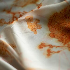 May include: A white fabric featuring a botanical print with orange and rust-colored leaf and plant silhouettes. The material has a soft, draped texture with folds that create light and shadow across the surface, highlighting the organic patterns.