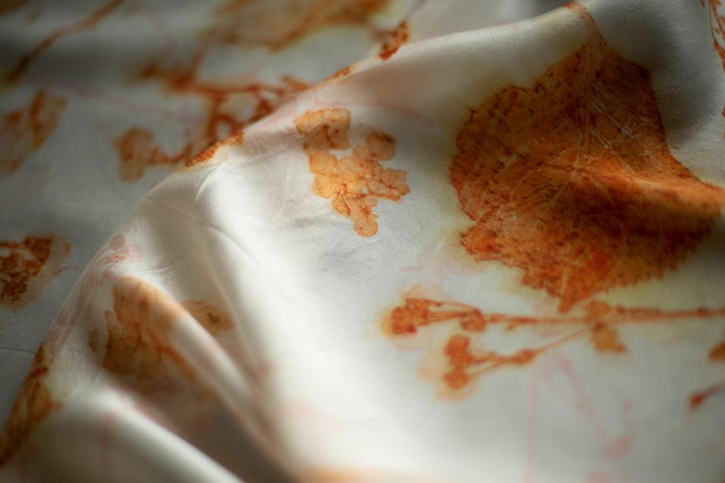 May include: A white fabric featuring a botanical print with orange and rust-colored leaf and plant silhouettes. The material has a soft, draped texture with folds that create light and shadow across the surface, highlighting the organic patterns.