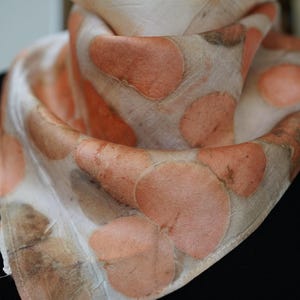 May include: A lightweight scarf featuring a botanical print with soft peach and muted brown circular leaf patterns on an off-white background. The fabric has a smooth, draped texture with visible hemmed edges, showcasing a natural, organic aesthetic.