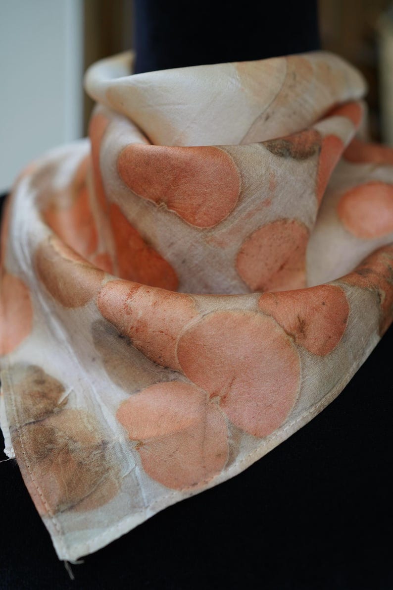 May include: A lightweight scarf featuring a botanical print with soft peach and muted brown circular leaf patterns on an off-white background. The fabric has a smooth, draped texture with visible hemmed edges, showcasing a natural, organic aesthetic.