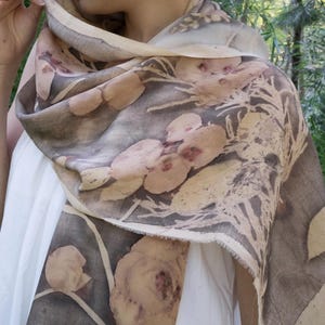 May include: A floral print scarf in shades of brown, beige, and gray. The scarf features a botanical design with large flower and leaf patterns. The fabric appears to be lightweight and draped over the shoulders.