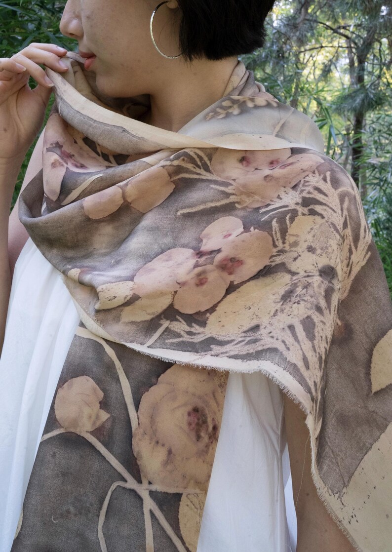May include: A floral print scarf in shades of brown, beige, and gray. The scarf features a botanical design with large flower and leaf patterns. The fabric appears to be lightweight and draped over the shoulders.