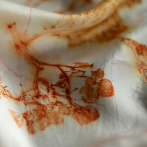 May include: A white fabric featuring a botanical print with rust-colored leaf and flower patterns. The design displays organic, detailed impressions of plant structures scattered across the smooth, folded surface of the textile material.