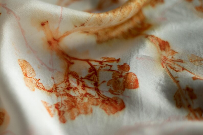 May include: A white fabric featuring a botanical print with rust-colored leaf and flower patterns. The design displays organic, detailed impressions of plant structures scattered across the smooth, folded surface of the textile material.
