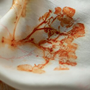 May include: A white fabric featuring a botanical print in shades of orange and rust. The design displays detailed floral and branch motifs with a soft, organic appearance. The material is folded, creating gentle curves and shadows across the surface.