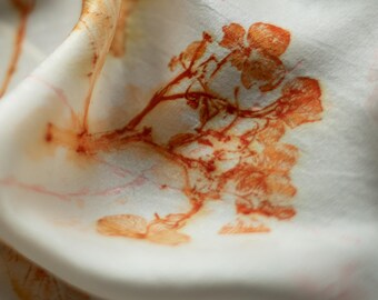 Puede incluir: Una tela blanca con un estampado botánico en tonos naranja y óxido. El diseño muestra motivos florales y de ramas detallados con un aspecto suave y orgánico. El material está plegado, creando curvas y sombras suaves en toda la superficie.