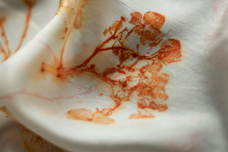 May include: A white fabric featuring a botanical print in shades of orange and rust. The design displays detailed floral and branch motifs with a soft, organic appearance. The material is folded, creating gentle curves and shadows across the surface.
