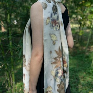 May include: A long, off-white rectangular scarf featuring a botanical print with various brown, yellow, and dark blue leaf and flower patterns. The fabric has a soft, draped appearance with raw, slightly frayed edges along the length of the textile.