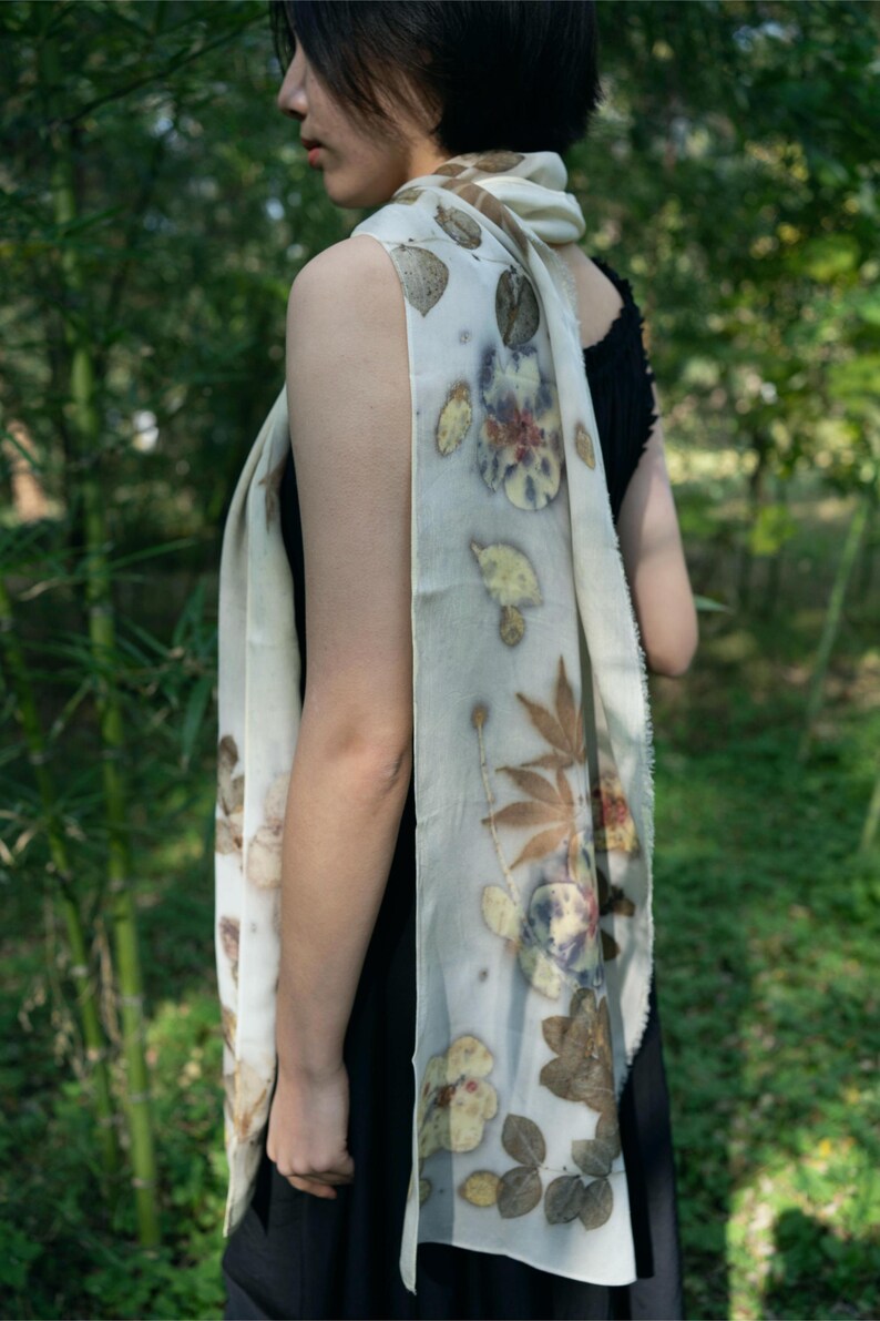 May include: A long, off-white rectangular scarf featuring a botanical print with various brown, yellow, and dark blue leaf and flower patterns. The fabric has a soft, draped appearance with raw, slightly frayed edges along the length of the textile.