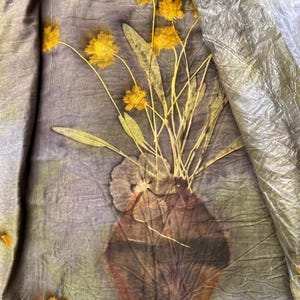 May include: A piece of fabric featuring a botanical print of yellow flowers in a brown vase. The textile has a crinkled texture with a muted grey-green background. A clear plastic sheet partially covers the upper right corner of the fabric.