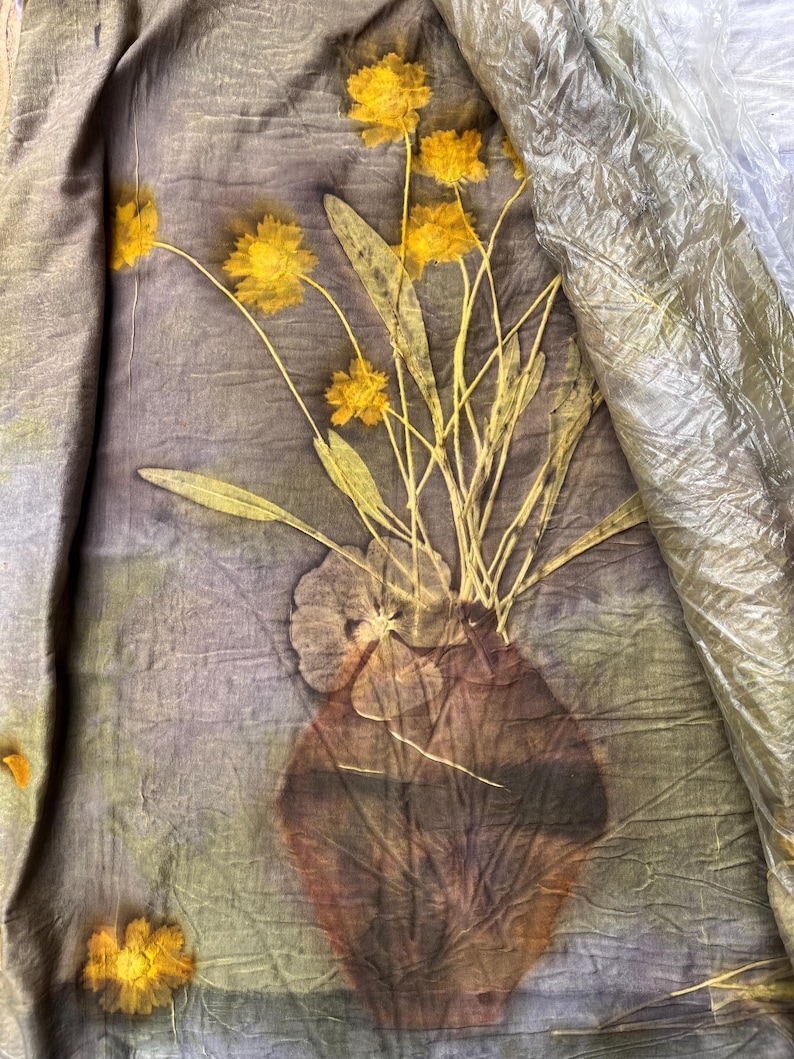 May include: A piece of fabric featuring a botanical print of yellow flowers in a brown vase. The textile has a crinkled texture with a muted grey-green background. A clear plastic sheet partially covers the upper right corner of the fabric.