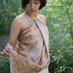 May include: A light peach-coloured halter top made from a soft, draped fabric featuring a botanical print of orange flowers and green leaves. The garment is secured with thin, light-coloured straps adorned with small, smooth, oval-shaped beads.
