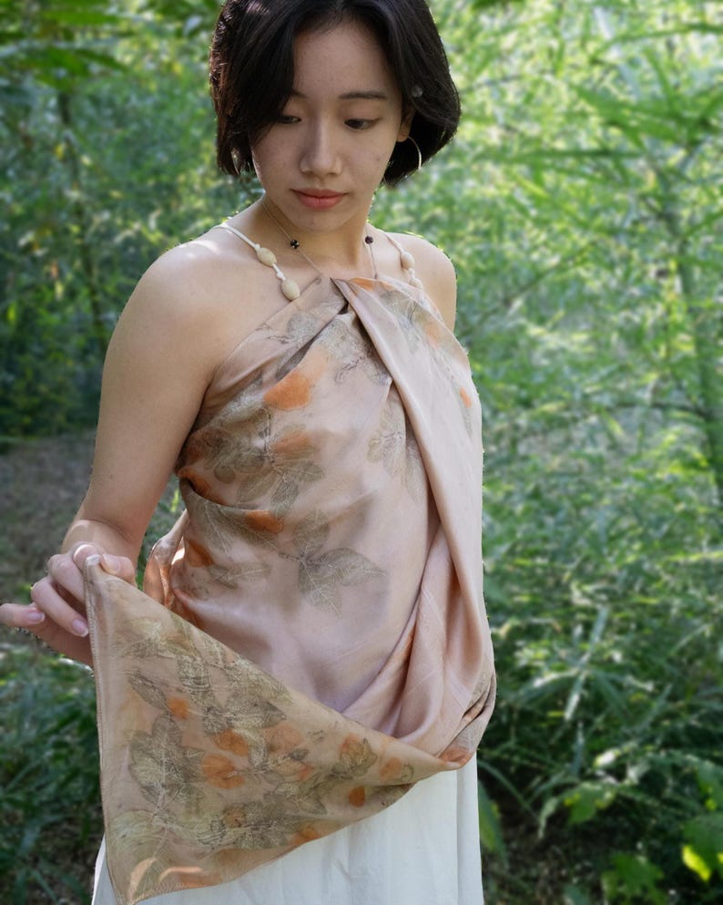 May include: A light peach-coloured halter top made from a soft, draped fabric featuring a botanical print of orange flowers and green leaves. The garment is secured with thin, light-coloured straps adorned with small, smooth, oval-shaped beads.