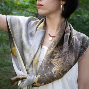 May include: A square silk scarf featuring a botanical print with shades of grey, charcoal, and muted yellow. The scarf is tied in a knot at the front, displaying a textured, organic pattern of leaves and branches across the fabric surface.