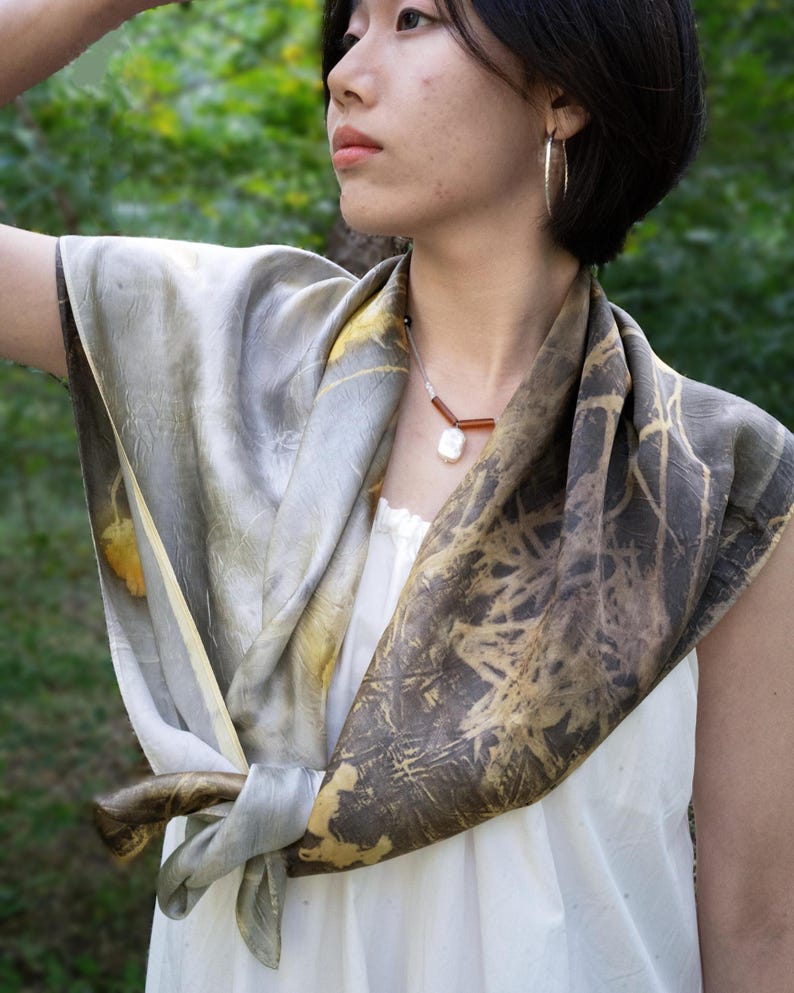 May include: A square silk scarf featuring a botanical print with shades of grey, charcoal, and muted yellow. The scarf is tied in a knot at the front, displaying a textured, organic pattern of leaves and branches across the fabric surface.