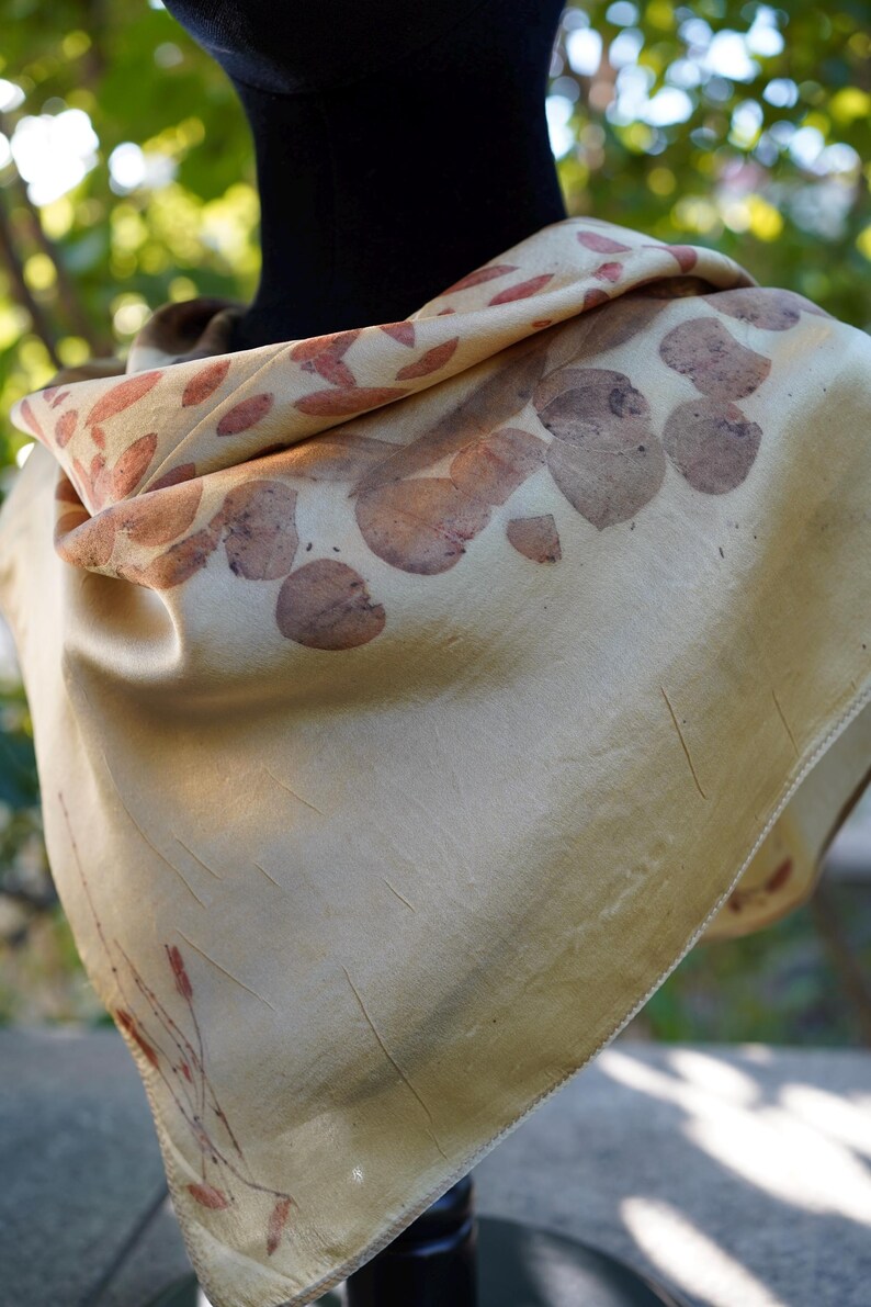 Pu&ograve; includere: Un foulard quadrato color crema chiaro caratterizzato da una stampa botanica con motivi di foglie e steli color bruno-rossastro sparsi. Il tessuto ha una finitura liscia e leggermente lucida con pieghe visibili e un orlo cucito.