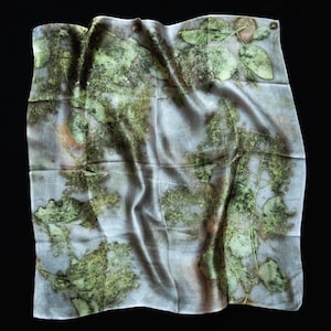 May include: A square scarf featuring a botanical print with various green leaf patterns and subtle brown accents on a light gray background. The fabric has a smooth, slightly reflective surface with natural folds and a soft, draped appearance.