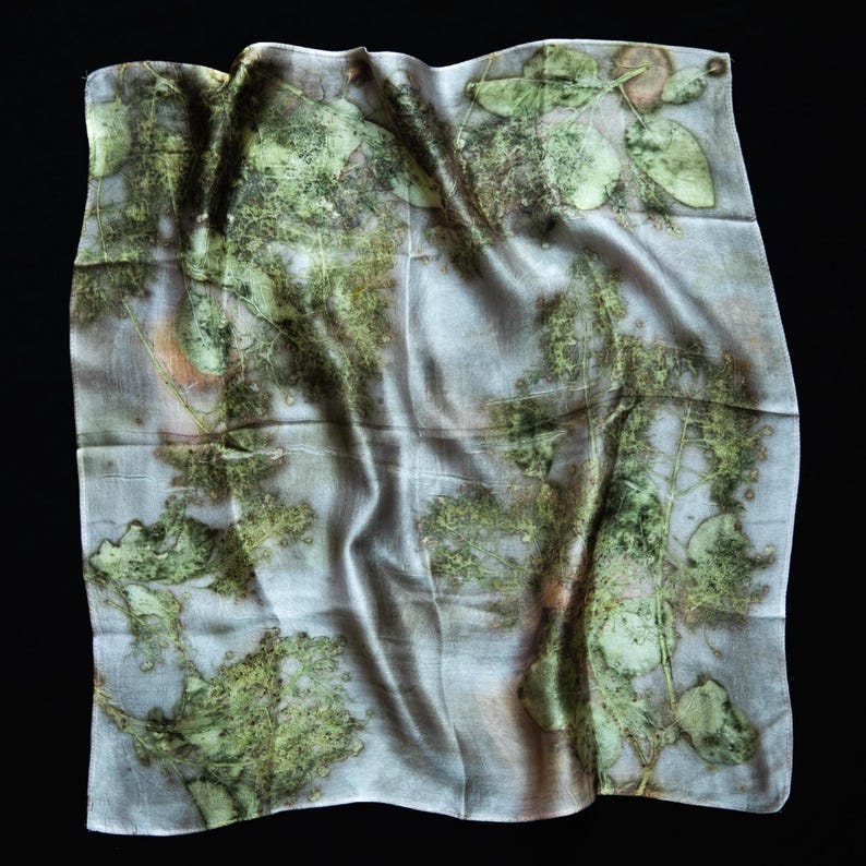 May include: A square scarf featuring a botanical print with various green leaf patterns and subtle brown accents on a light gray background. The fabric has a smooth, slightly reflective surface with natural folds and a soft, draped appearance.