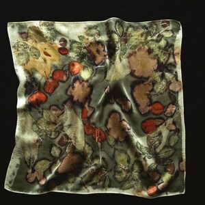 May include: A square silk scarf featuring a botanical print with various green leaves and reddish-orange berries scattered across a muted sage green background. The fabric has a soft, draped texture with visible natural plant imprints throughout.
