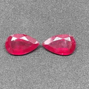 May include: Two faceted, pear-shaped rubies. The gemstones are a deep, vibrant red color and have a polished, reflective surface. The gems are displayed against a dark gray background.
