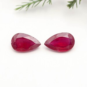 Pode incluir: Dois rubis lapidados em formato de pera com uma cor vermelha profunda e vibrante. Cada pedra preciosa apresenta uma extremidade pontiaguda e uma base arredondada, com superfície polida e inclusões internas visíveis na estrutura vermelha.