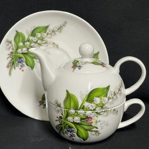 May include: A white porcelain tea set comprising a teapot, teacup, and saucer. The set is adorned with a floral design of white lily of the valley flowers, green leaves, and small blue and pink details. The teapot has a lid and two handles.