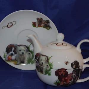 Pode incluir: Conjunto de chá de cerâmica branca com tema de cães. O bule e a xícara apresentam diferentes raças de cães, incluindo um dachshund, um cocker spaniel e um terrier. O pires tem um design de ferradura e uma borda de grama verde.