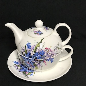 Könnte beinhalten: Ein weißes Porzellan-Teekannen- und Teetassen-Set mit einem Blumendesign mit blauen, rosa und weißen Blumen. Die Teekanne hat einen Deckel und einen Griff, und die Teetasse hat einen Griff. Das Set befindet sich auf einer weißen Untertasse.