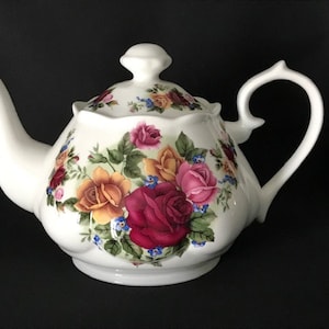 May include: White porcelain teapot with a floral design featuring pink, red, yellow, and blue roses. The teapot has a scalloped edge and a curved handle.