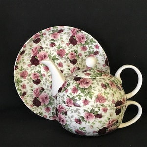 Puede incluir: Un juego de tetera y platillo de porcelana blanca con un patrón floral de rosas rosas y burdeos. La tetera tiene una tapa y un asa, y el platillo es redondo.