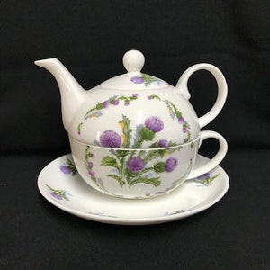May include: A white porcelain teapot and teacup set with a floral design featuring purple thistles. The set is decorated with green leaves and a white background.