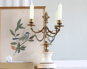 May include: A gold metal candelabra with three candle holders and a white marble base. The candelabra has a chain detail and is decorated with ornate details. The candelabra is in front of a framed print of a bird on a branch.