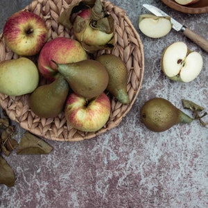 Op de afbeelding: Een rieten mand gevuld met groene peren en rode appels. De mand staat op een rustiek bruin oppervlak met wat bladeren eromheen.