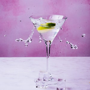 May include: A martini glass filled with a clear liquid and a lime wedge, with water splashing around it. There are three ice cubes on the table.