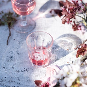 Op de afbeelding: Twee heldere glazen bekers gevuld met roze vloeistof, die op een wit kantkleed staan.  Roze en witte bloemen zijn rond de bekers verspreid.