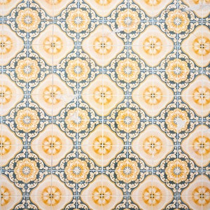 May include: Close-up of a tiled surface with a repeating pattern. The tiles feature a cream-colored background with yellow and blue floral designs. The pattern is symmetrical and creates a visually appealing texture.