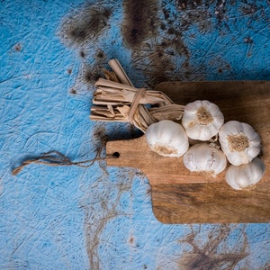 Op de afbeelding: Een bos knoflookbollen die met touw aan elkaar gebonden zijn en op een houten snijplank liggen. De plank ligt op een blauw en bruin getextureerd oppervlak.