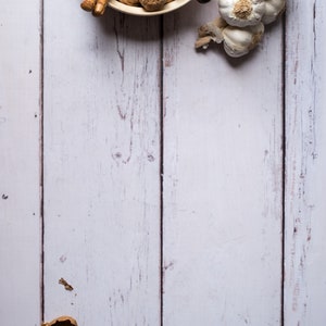 Op de afbeelding: Een witte kom gevuld met walnoten en een notenkraker. De kom staat op een wit houten oppervlak. Er zijn ook verschillende teentjes knoflook aan de rechterkant van de afbeelding.
