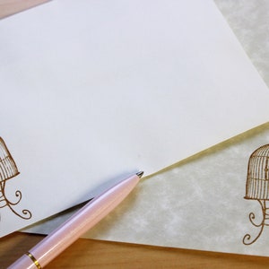 May include: A white envelope with a brown birdcage design on the bottom left corner. A pink pen is lying on top of the envelope. The envelope is on a wooden surface.