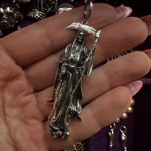 May include: A silver pendant depicting the Grim Reaper holding a scythe. The pendant is detailed with a skeletal face and flowing robes. Other silver pendants and jewelry are visible in the background.