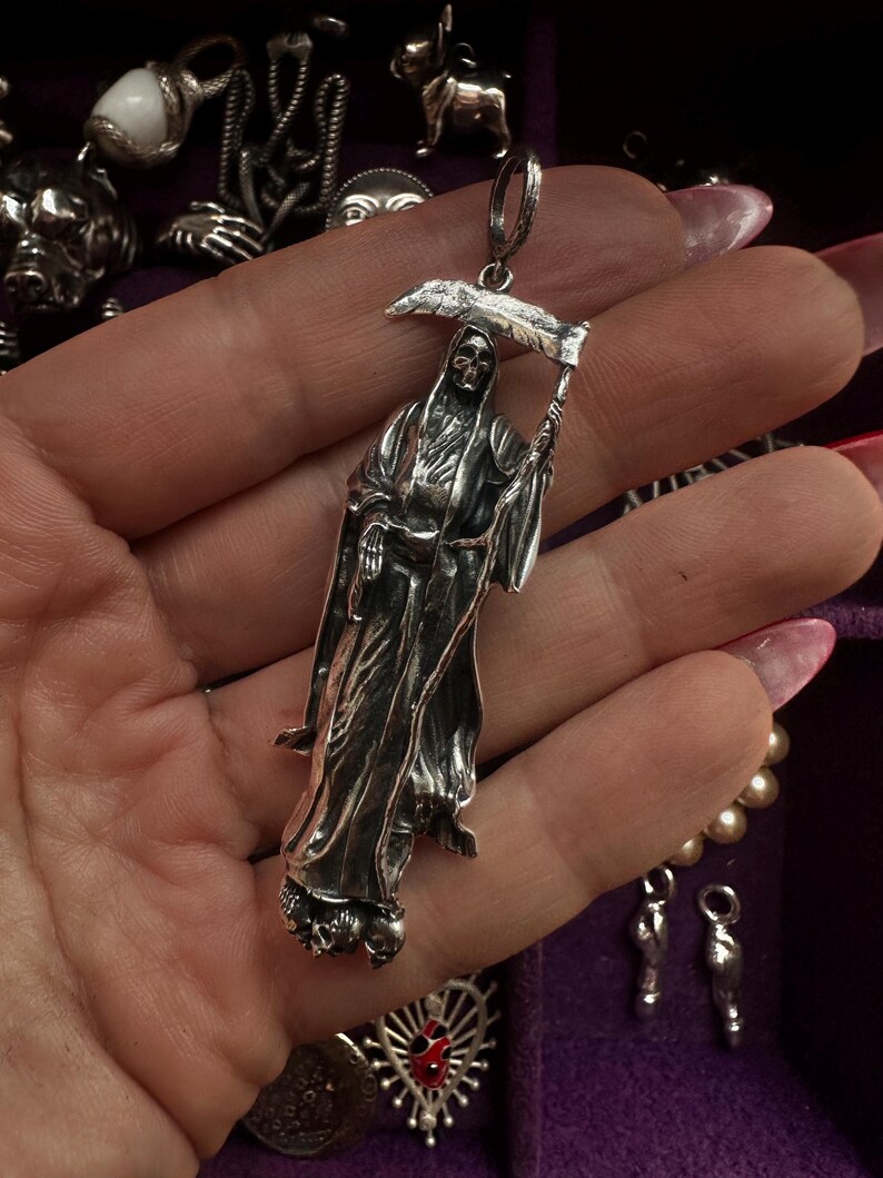 May include: A silver pendant depicting the Grim Reaper holding a scythe. The pendant is detailed with a skeletal face and flowing robes. Other silver pendants and jewelry are visible in the background.