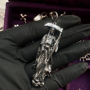 May include: A silver pendant depicting the Grim Reaper holding a scythe. The pendant is detailed with a skeletal face and flowing robes. The Reaper stands atop a base of skulls. The pendant is held by a black gloved hand.