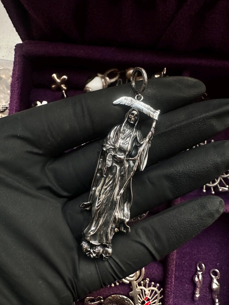 May include: A silver pendant depicting the Grim Reaper holding a scythe. The pendant is detailed with a skeletal face and flowing robes. The Reaper stands atop a base of skulls. The pendant is held by a black gloved hand.