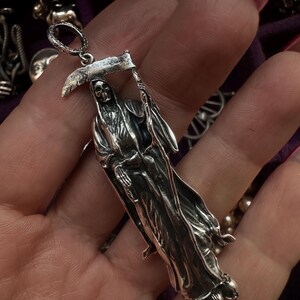 May include: A silver pendant depicting the Grim Reaper holding a scythe. The pendant is detailed with a skeletal face and flowing robes. The bail is a small loop for a chain. The pendant is likely made of sterling silver.