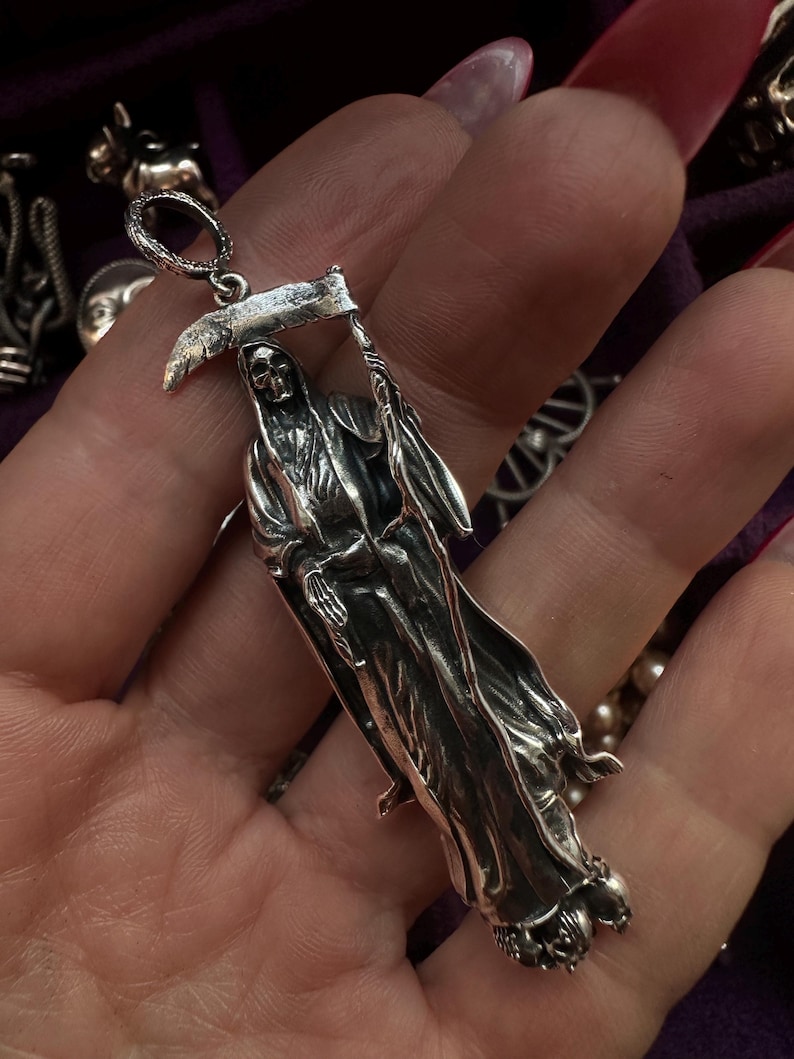 May include: A silver pendant depicting the Grim Reaper holding a scythe. The pendant is detailed with a skeletal face and flowing robes. The bail is a small loop for a chain. The pendant is likely made of sterling silver.