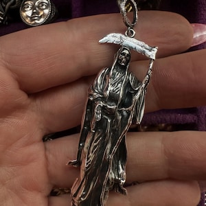 May include: A silver pendant depicting the Grim Reaper holding a scythe. The pendant is detailed with a skeletal face and flowing robes. Other silver jewelry pieces are visible in the background, including a ring with a moon face.