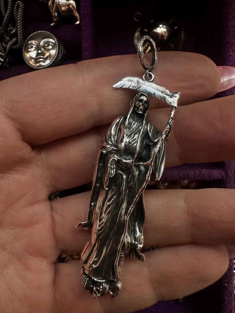 May include: A silver pendant depicting the Grim Reaper holding a scythe. The pendant is detailed with a skeletal face and flowing robes. Other silver jewelry pieces are visible in the background, including a ring with a moon face.