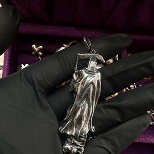 May include: A silver pendant in the shape of the Grim Reaper, holding a scythe. The pendant has intricate details in the flowing robes and hood. The pendant is held against a purple background.