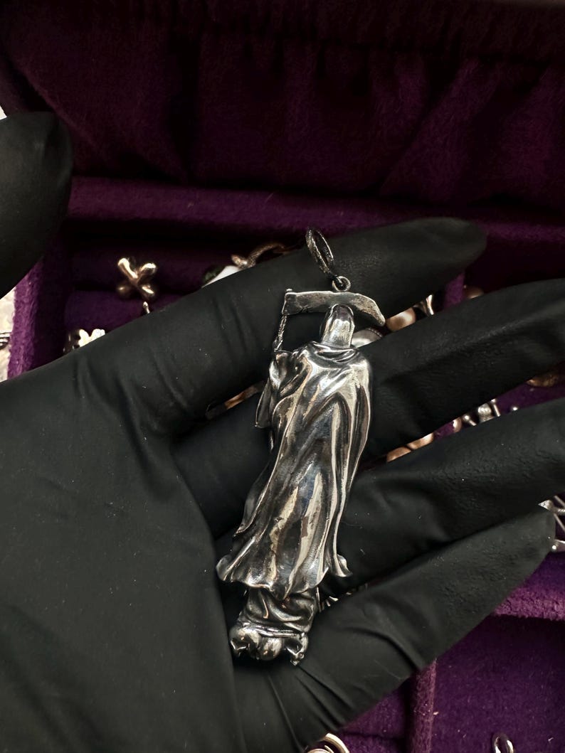 May include: A silver pendant in the shape of the Grim Reaper, holding a scythe. The pendant has intricate details in the flowing robes and hood. The pendant is held against a purple background.